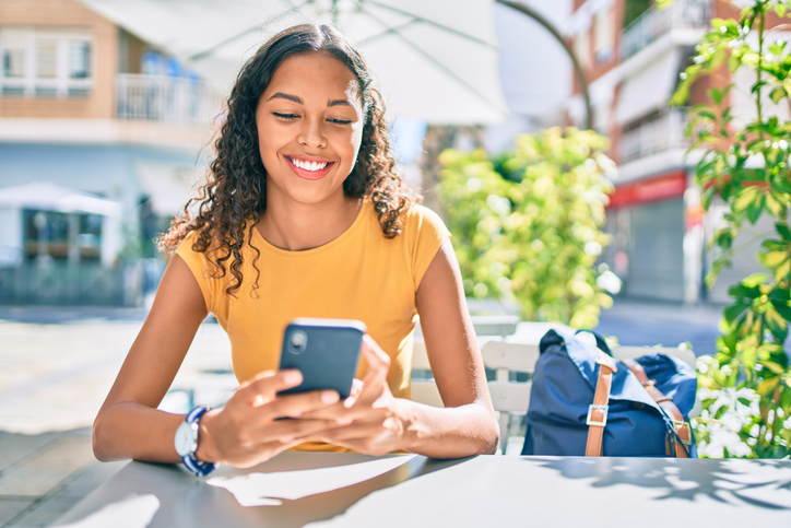 Happy young woman sitting outside with her backpack and checking her text messages.