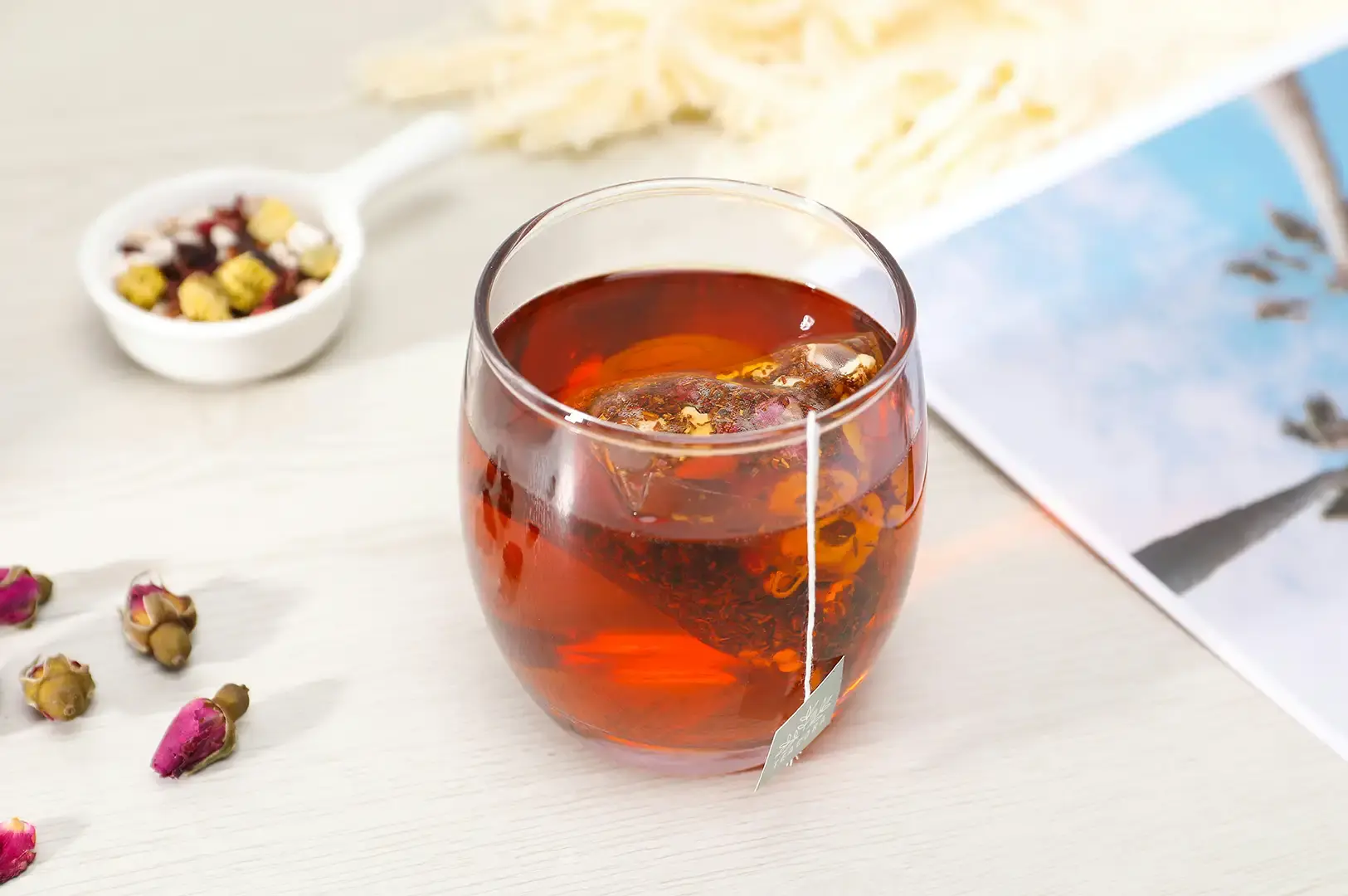 A clear glass mug containing warm amber tea and a steeping teabag sits on a light wooden table alongside scattered dried pink rosebuds and a white ceramic scoop filled with mixed herbal tea ingredients. A relaxed, bright aesthetic is enhanced by a postcard displaying a sunny sky in the background.