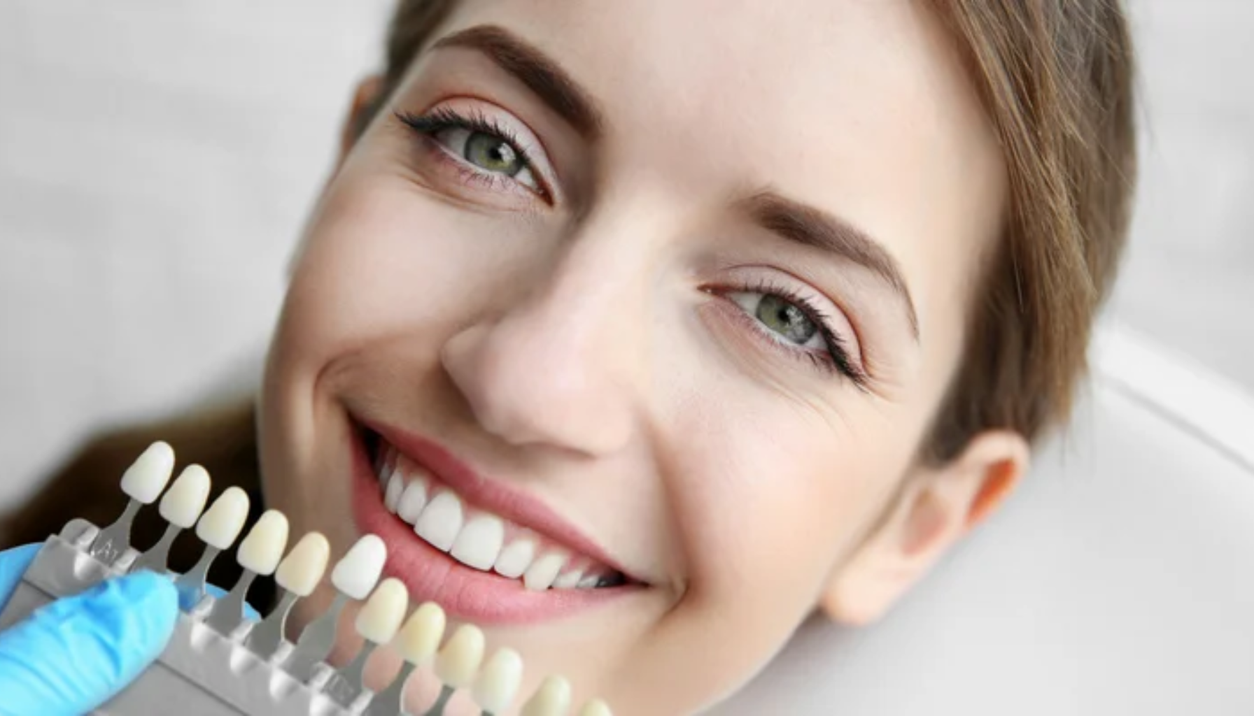 "Kvinne smiler i tannlegestol mens tannlege sammenligner tannfarger med fargeskala for bleking eller fasetter."