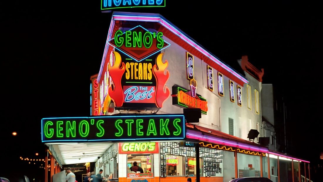 Brightly lit storefront of Geno's Steaks at night, featuring bold neon signs in red, yellow, green, and blue promoting cheesesteaks, fries, and hoagies, with flames and lettering that say โThe Bestโ above the entrance.
