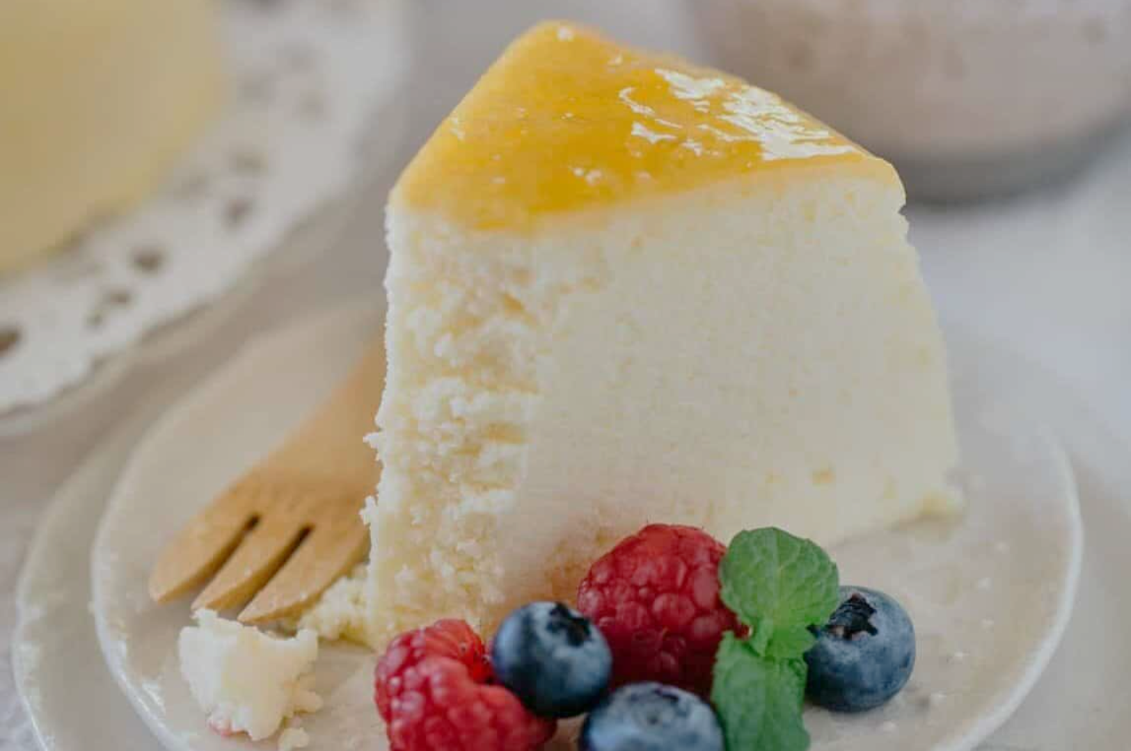 A slice of fluffy cheesecake with a glossy yellow glaze is on a white plate. It is garnished with raspberries, blueberries, and mint leaves, next to a wooden fork.