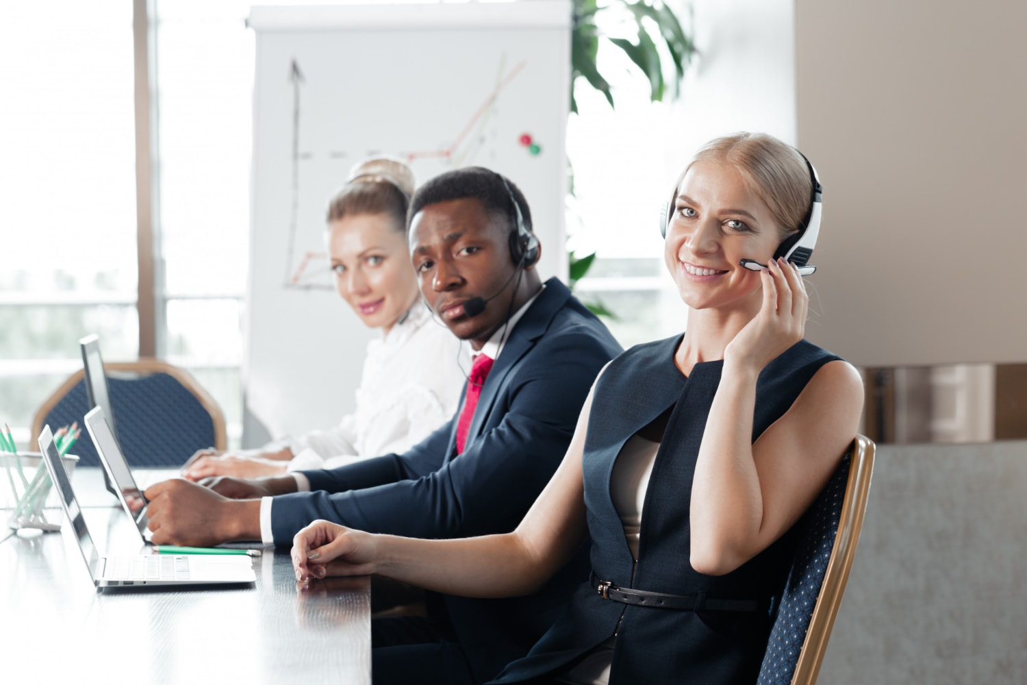 Three call center agents in formal attire sit at a polished wooden table, wearing headsets and working on laptops. The presence of data charts in the background emphasizes the role of real-time contact center analytics in improving customer interactions.