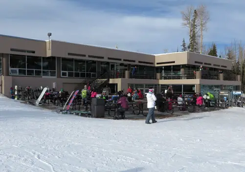 The image shows the exterior of Navajo Lodge, surrounded by snow-capped mountains, capturing the beauty of winter in Brian Head, Utah. The lodge is a welcoming spot for families and friends looking to enjoy delicious menu items such as grilled chicken sandwiches and burgers after a day on the slopes.