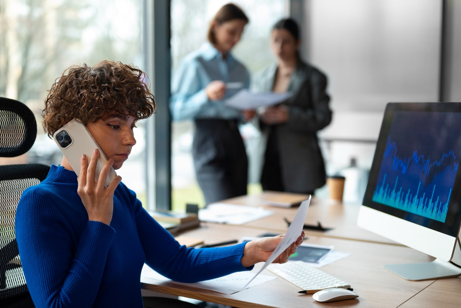 A businesswoman in a blue sweater talking on a smartphone while reviewing documents at her desk, with a computer screen displaying financial graphs. This reflects the power of Business phone solutions and Cloud communications in managing corporate operations efficiently.