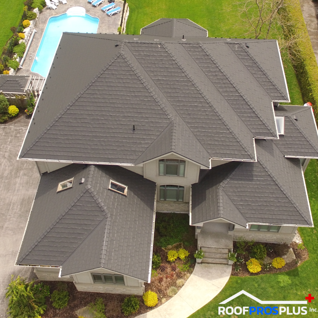 An aerial view of a large house with a dark grey metal roof. A lush green lawn and a swimming pool with lounge chairs are behind the house.