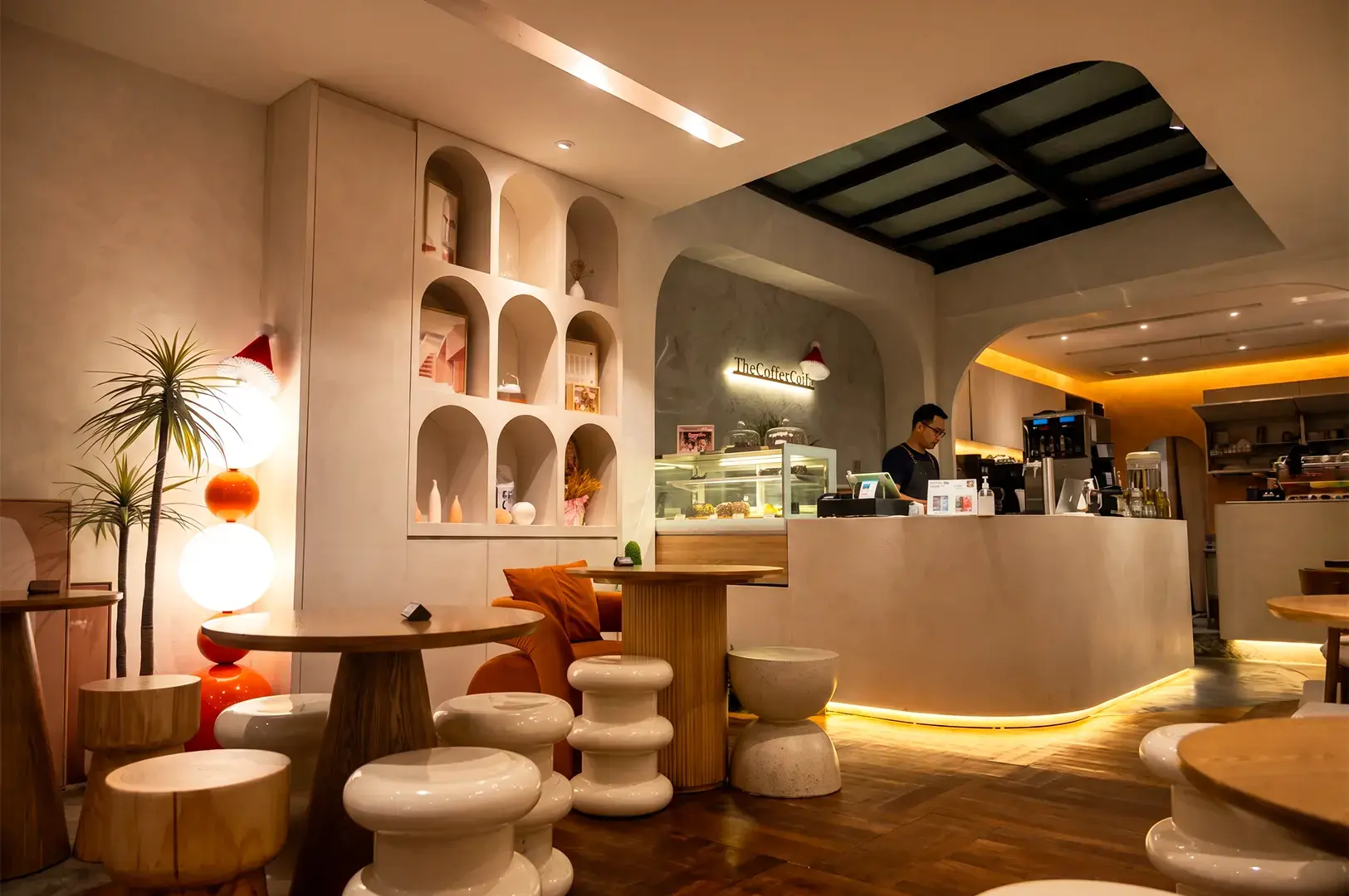 Interior view of Coffee Code, with empty chairs and wooden tables surrounding a counter with a staff working behind, its white interior lit by warm yellow lights.