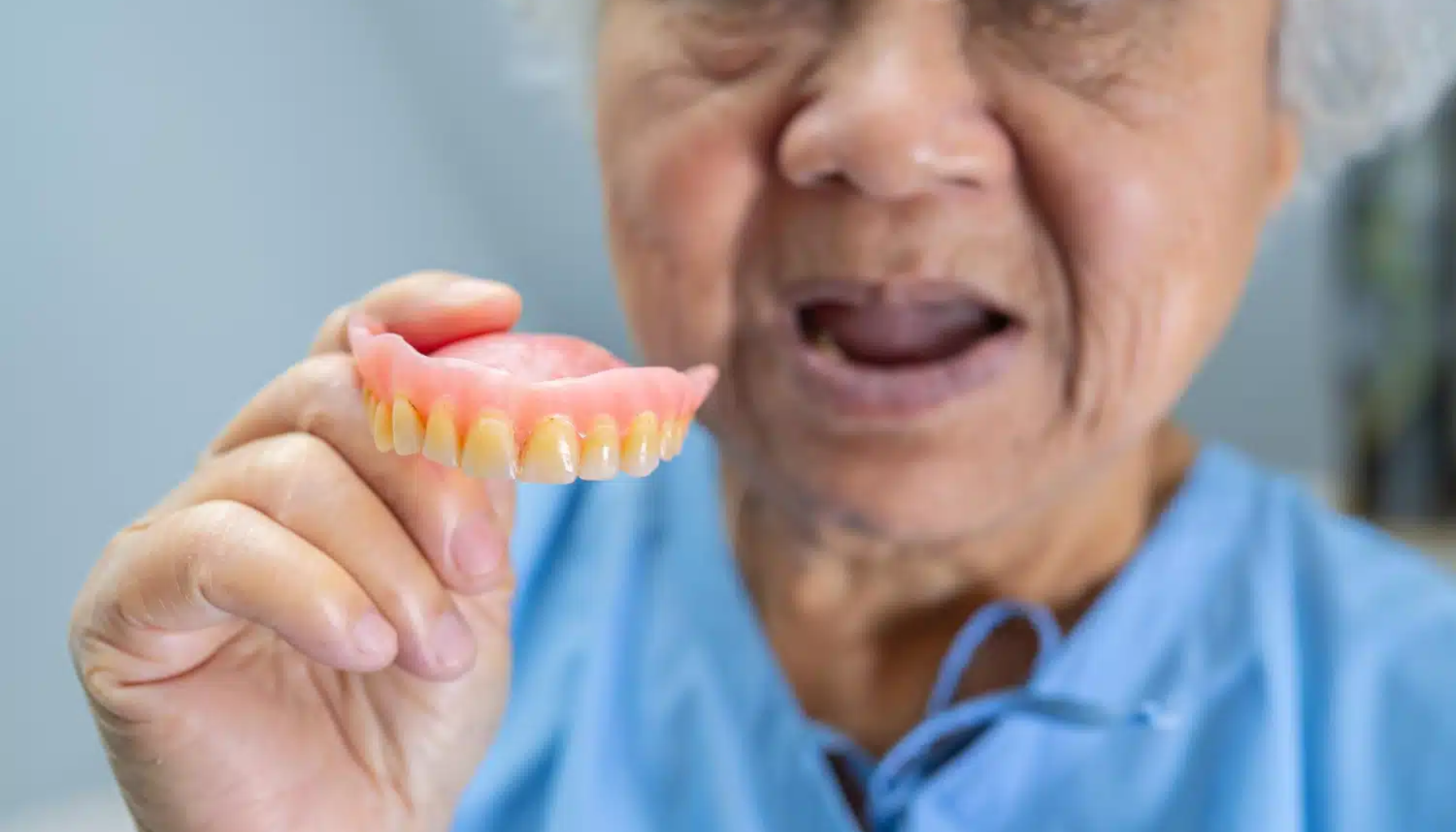 Gebiss – En komplett guide til avtakbare tannproteser 19 Eldre kvinne i blå skjorte holder et sett med gebiss i hånden.