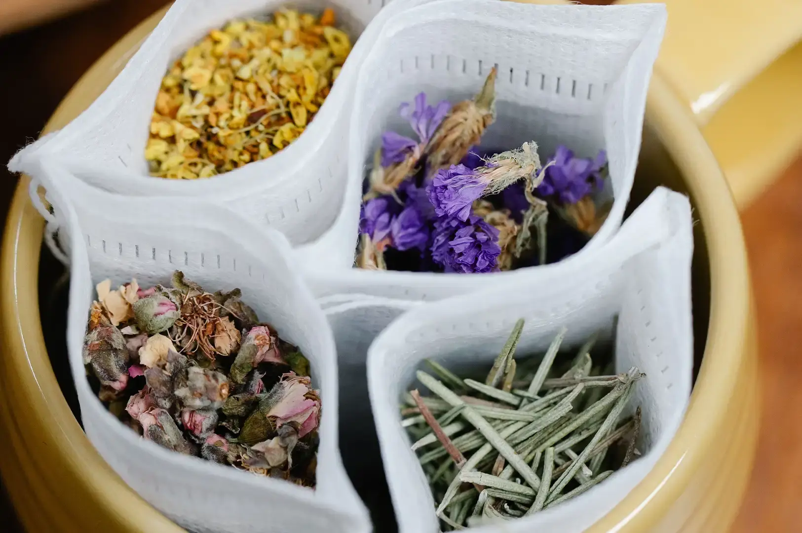 A top-down view looking into a ceramic container filled with four open white mesh tea bags, each neatly packed with distinct dried botanicals: yellow osmanthus flowers, bright purple petals, dried rosebuds, and needle-like green pine or rosemary leaves.