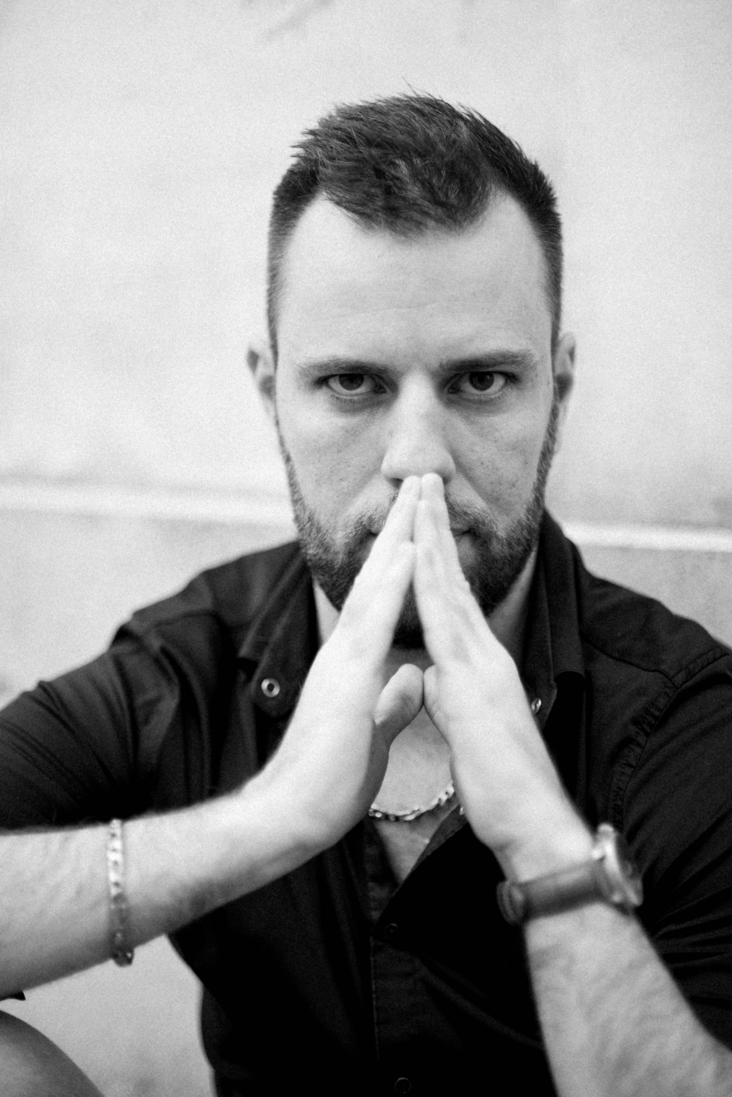 Black-and-white portrait of a bearded man in a dark button-up shirt, hands pressed together in front of his face in a focused, deliberate gesture — conveying patience, discipline, and quiet intensity.