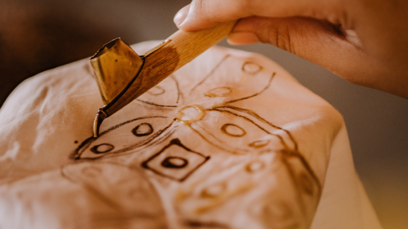 A person is using a wooden stick to draw on a piece of batik fabric.