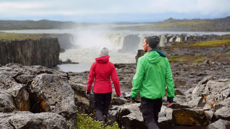 Two hikers in red and green jackets approach a wide waterfall.