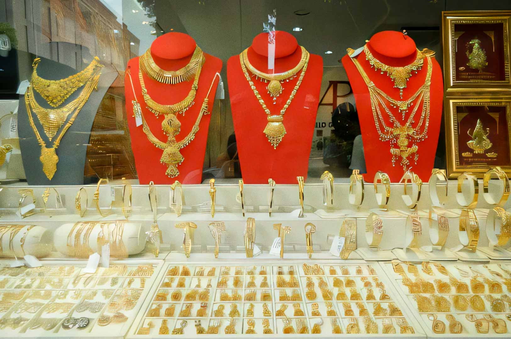 A jewelry shop display filled with gold necklaces, bangles, and rings arranged on red stands and trays behind a glass window in Little India.