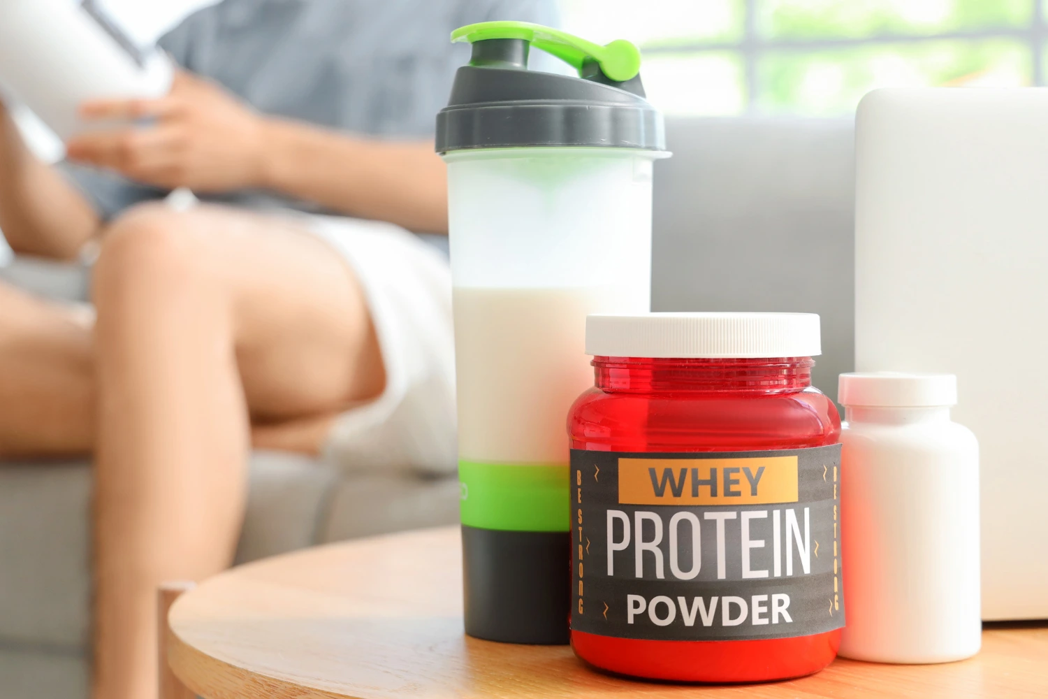 A shaker bottle, a jar of whey protein powder, and a supplement bottle sit on a table.