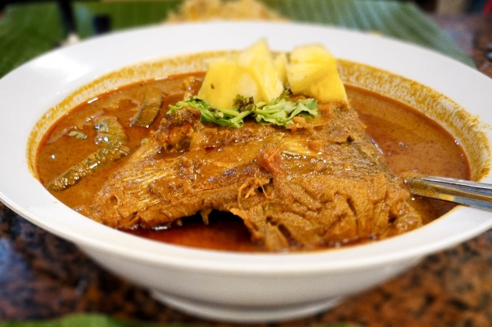 A bowl of fish head curry filled with a rich, reddish-brown gravy and topped with pineapple chunks and herbs.