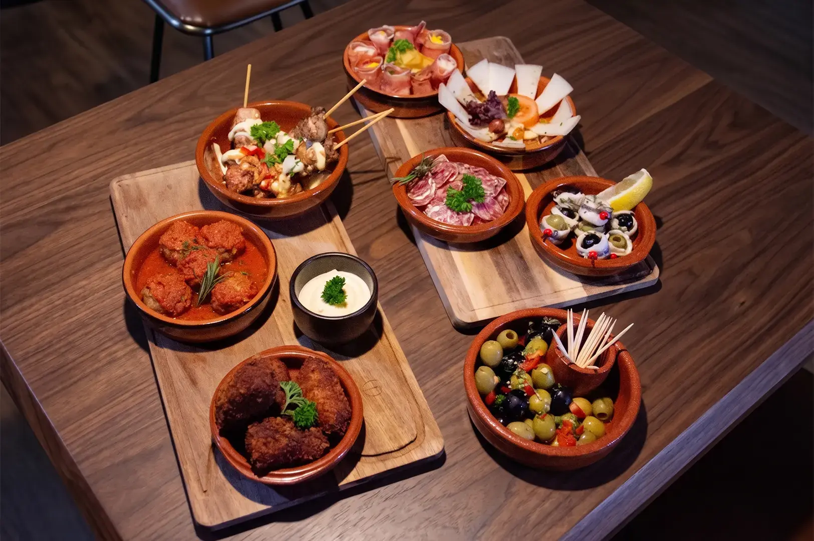 Assorted Spanish tapas served in clay bowls on wooden boards, featuring meat skewers, meatballs, olives, sliced cured meats, and fried appetizers.