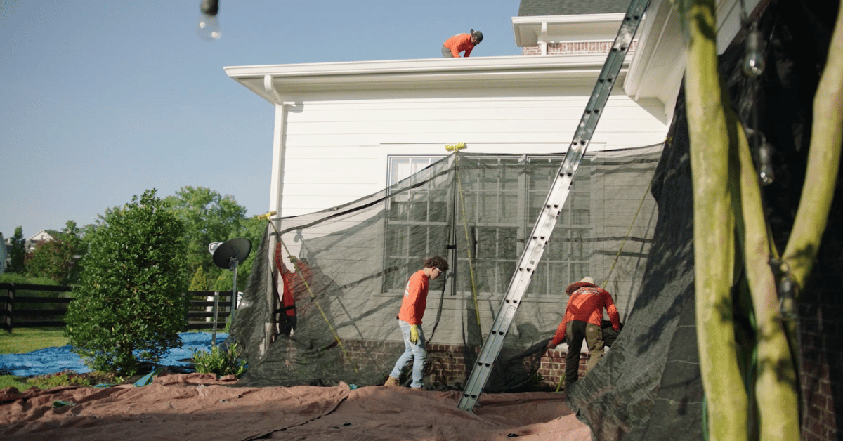 Roofing team installs protective mesh netting and ladder access along the side of a house to safeguard windows, siding, and landscaping during roofing work.