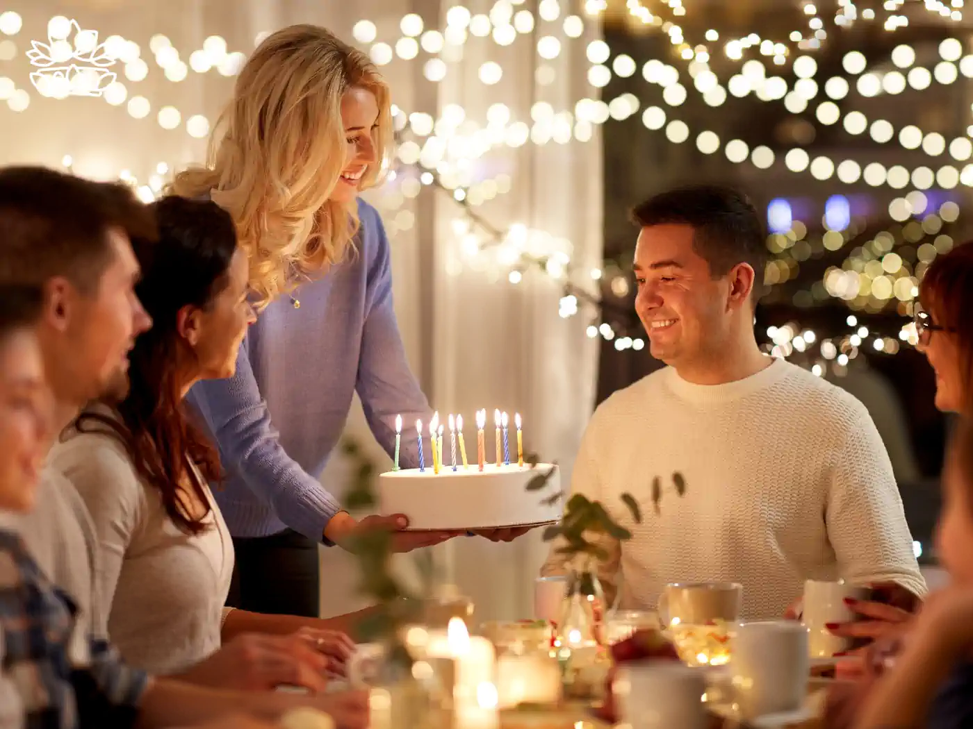 A woman presenting a birthday cake to a smiling man surrounded by friends in a warmly lit room. Fabulous Flowers and Gifts.