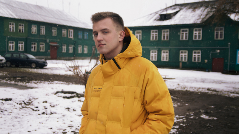 A person in a yellow gorpcore jacket stands before a green building on snow.