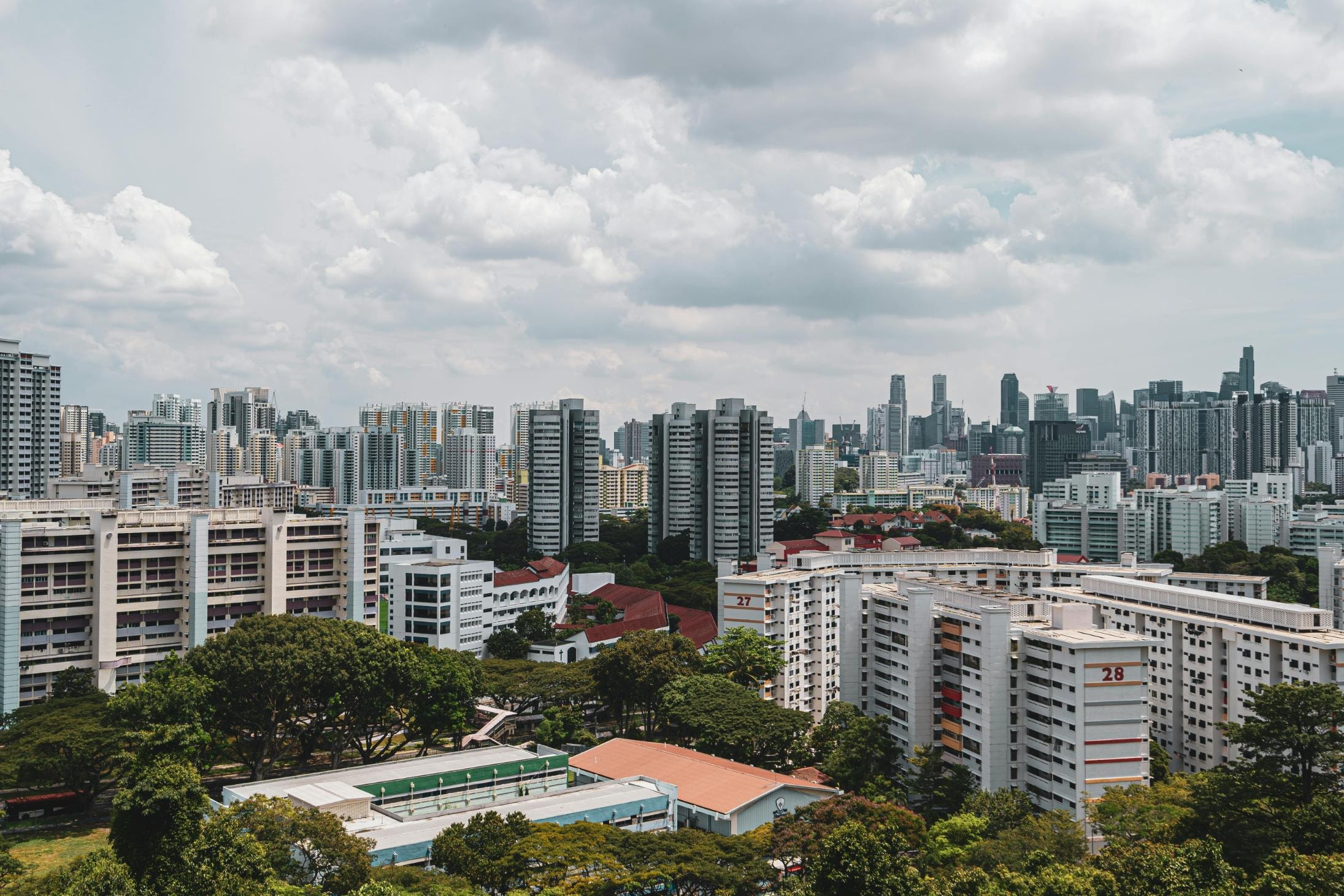 A dense urban landscape features clusters of white high-rise apartment blocks, some marked with large red numbers, nestled among lush green trees. In the background, a modern city skyline of skyscrapers rises against a dramatic sky filled with billowing white clouds.