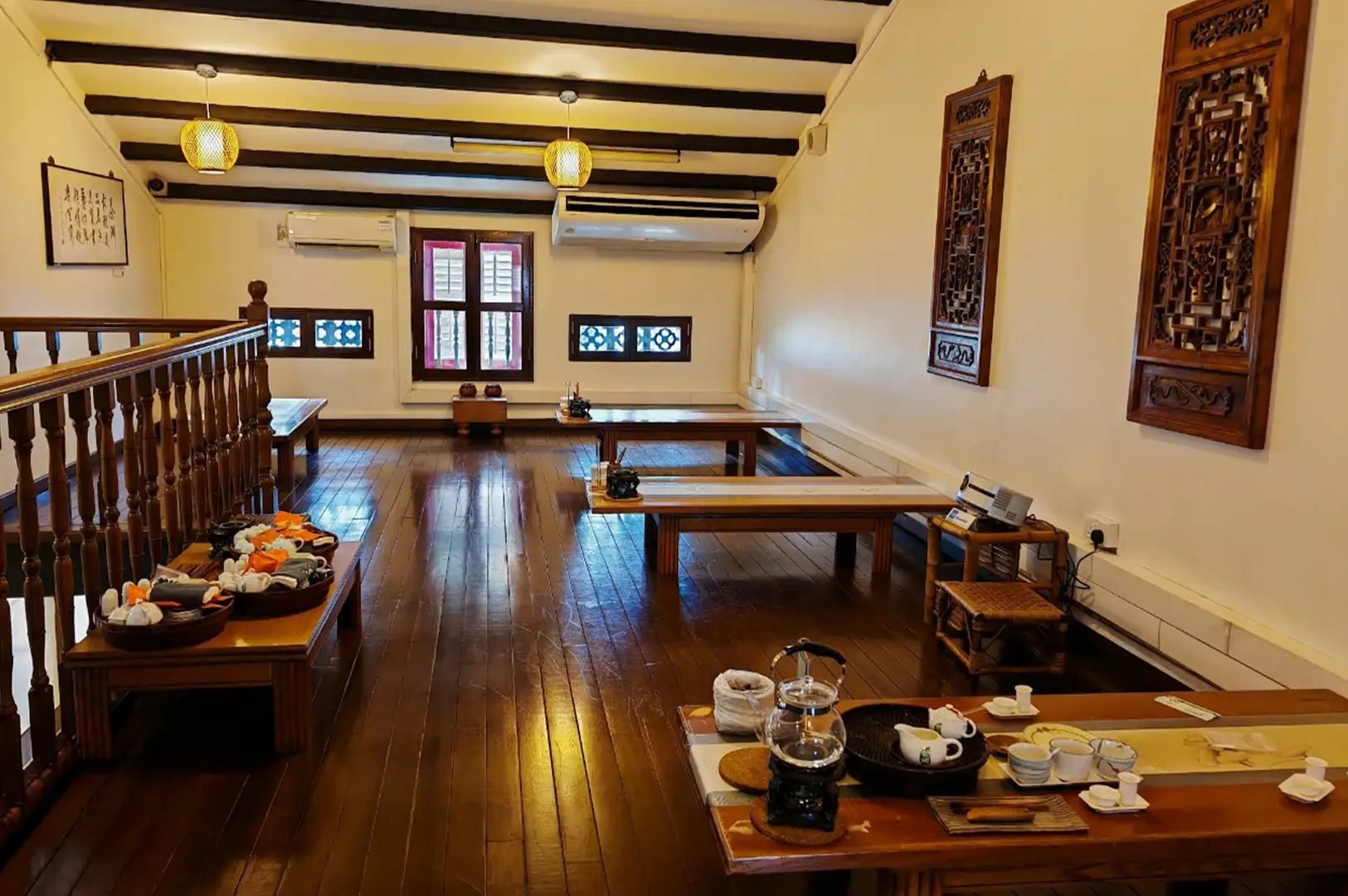 The interior of a spacious, traditional tea house features polished dark wooden floors and multiple low wooden tables arranged throughout the room for seated tea service. Decorative wooden panels adorn the white walls, and warm light shines down from paper lanterns hanging from the dark-beamed ceiling. On the tables in the foreground and near the railings, various tea sets, kettles, and small cups are neatly laid out, creating an inviting and tranquil atmosphere for guests to enjoy a tea experience.