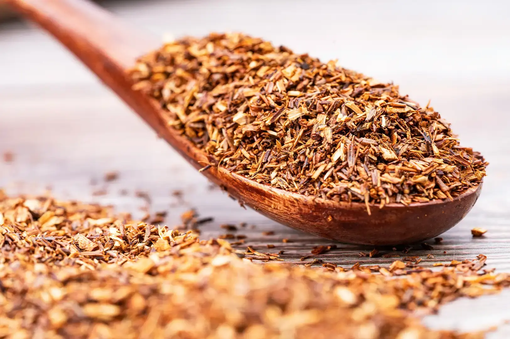 A close-up photograph highlights a dark wooden spoon filled to overflowing with fine, needle-like reddish-brown loose rooibos tea leaves. Additional loose tea leaves are strewn across the textured wooden surface below, emphasizing the dry, coarse texture of the tea blend.