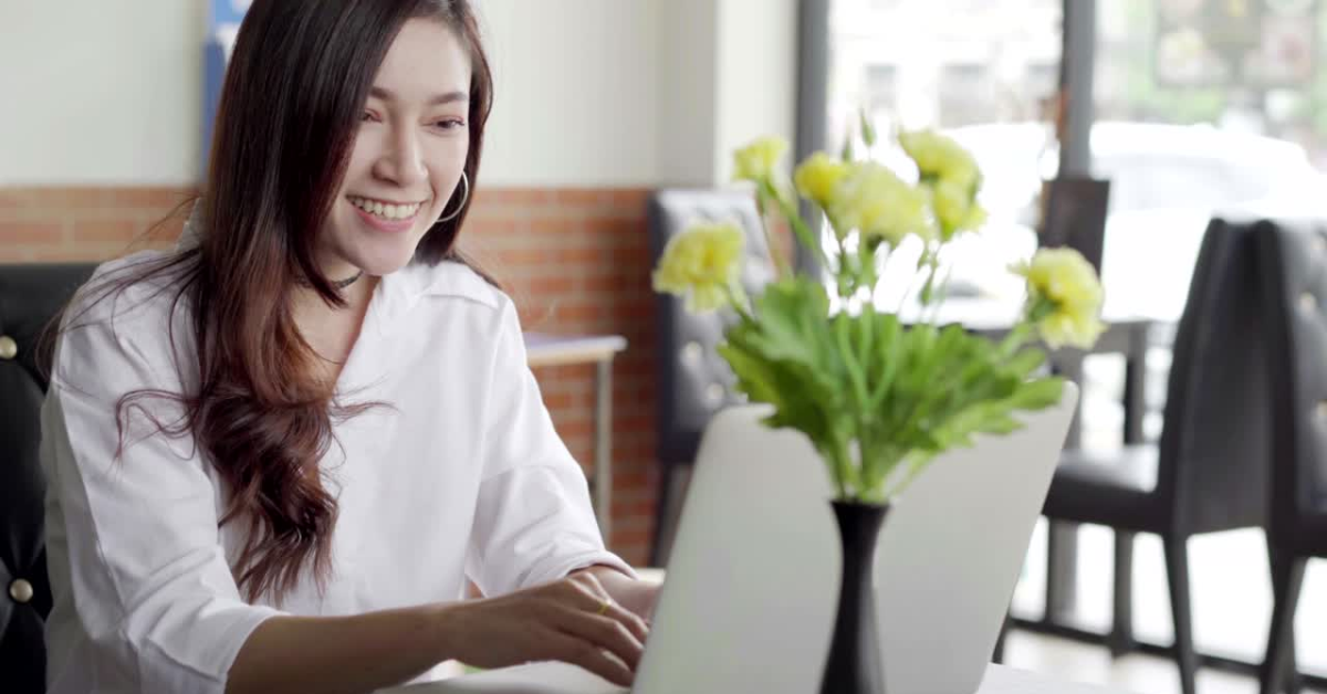 A smiling woman in a café working on her laptop, ensuring accuracy in completing her schedule SE tax form.