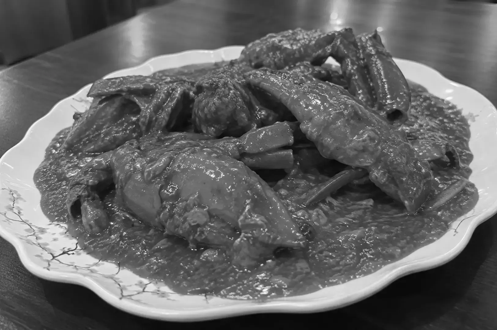 Whole chili crab with large claws and legs arranged on a white scalloped plate decorated with delicate floral patterns, covered in thick sauce and photographed from a low side angle.