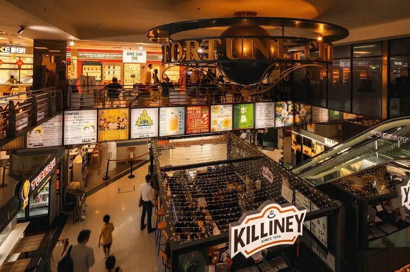 A bustling interior view of Fortune Centre showing multiple levels of brightly lit eateries, diners seated at tables, and a prominent decorative "Fortune Centre" sign hanging from the ceiling above.