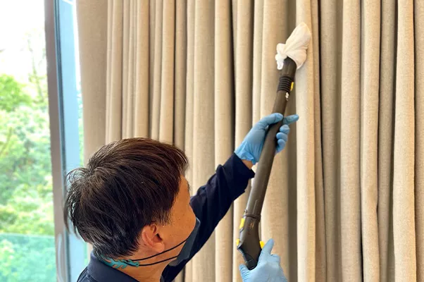 A cleaner wearing gloves and a mask uses a vacuum cleaner with a cloth attachment to clean tall beige curtains next to a large window with a green outdoor view.