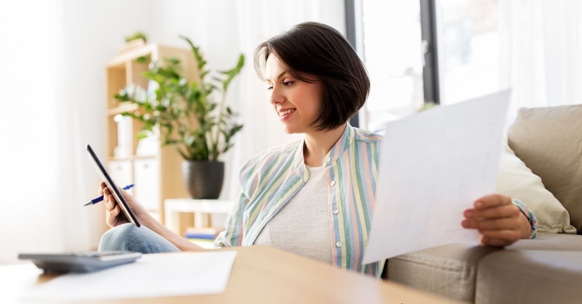 A woman reviewing her finances on a tablet while holding paperwork, likely tracking her payment report.