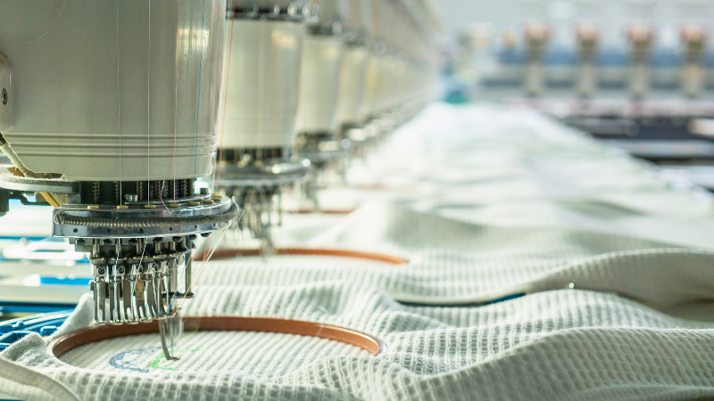 A close-up view of industrial embroidery machines working on white virgin polyester fabric in a factory setting.