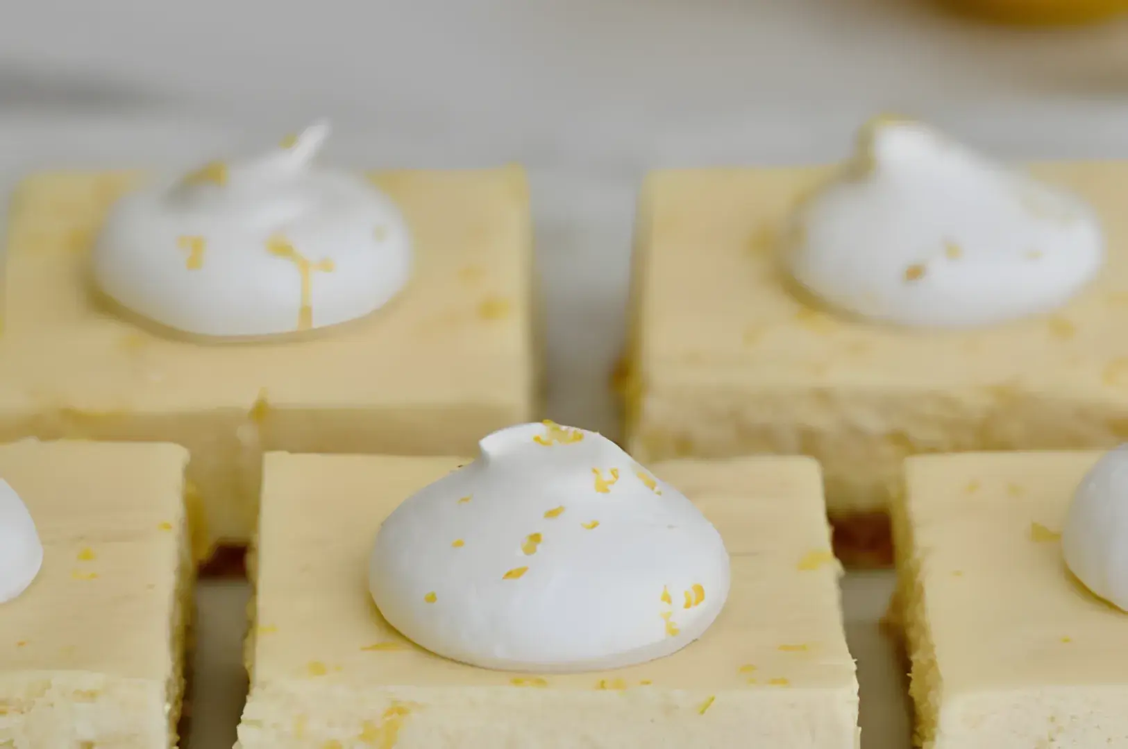 Close-up of creamy lemon bars topped with whipped cream and lemon zest. The smooth, pale yellow squares evoke a fresh and indulgent dessert feel.