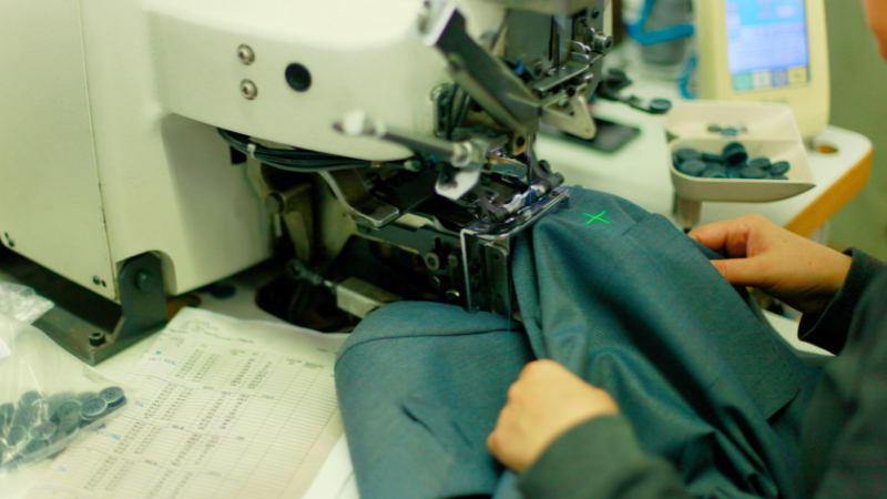 Close-up of a garment being sewn on a machine, with a tray of buttons nearby and a sheet of paper with columns of numbers.