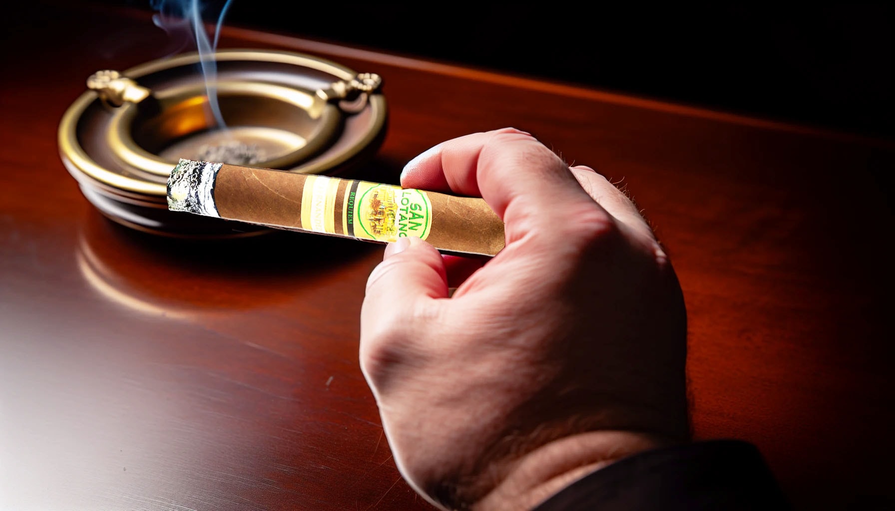 A hand holding a San Lotano Requiem Habano Robusto cigar with an ashtray in the background