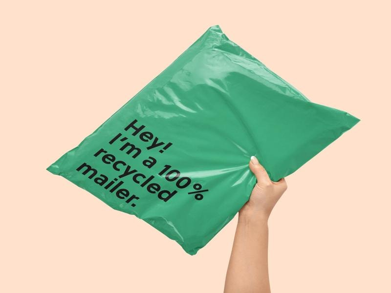 A person holds a bright green mailing bag against a soft beige background. The mailer is large, slightly crinkled, and made of recycled material. Bold black text on the bag reads, "Hey! I’m a 100% recycled mailer." The image promotes sustainable packaging, encouraging businesses and shoppers to choose environmentally friendly shipping materials.