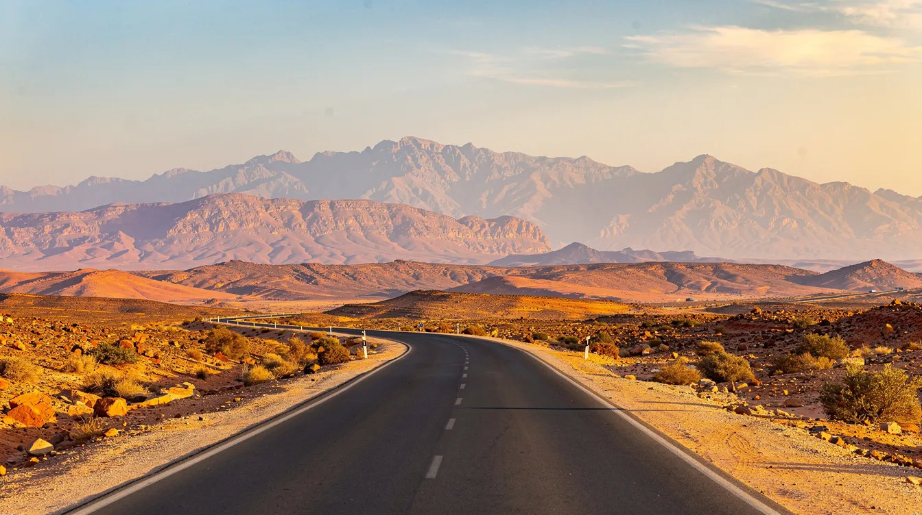 Una carretera panorámica se extiende en el sur de Marruecos, con impresionantes montañas del Atlas al fondo, creando un paisaje natural espectacular. Este entorno es ideal para disfrutar de un viaje en coche y explorar la belleza del país.