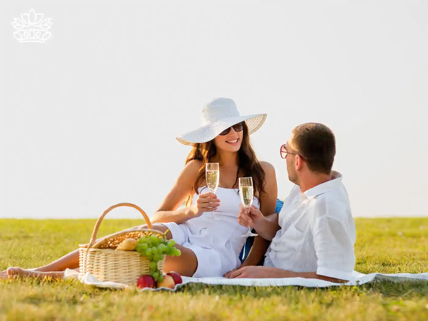  A couple enjoying a romantic picnic with a basket of grapes, apples, and champagne, sitting on a white blanket. Fabulous Flowers and Gifts - Picnic Gift Baskets Collection.