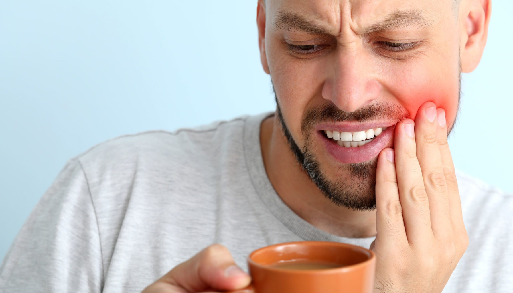 Mann med tannverk holder seg til kinnet og reagerer på smerte ved å drikke varm drikke fra kopp.