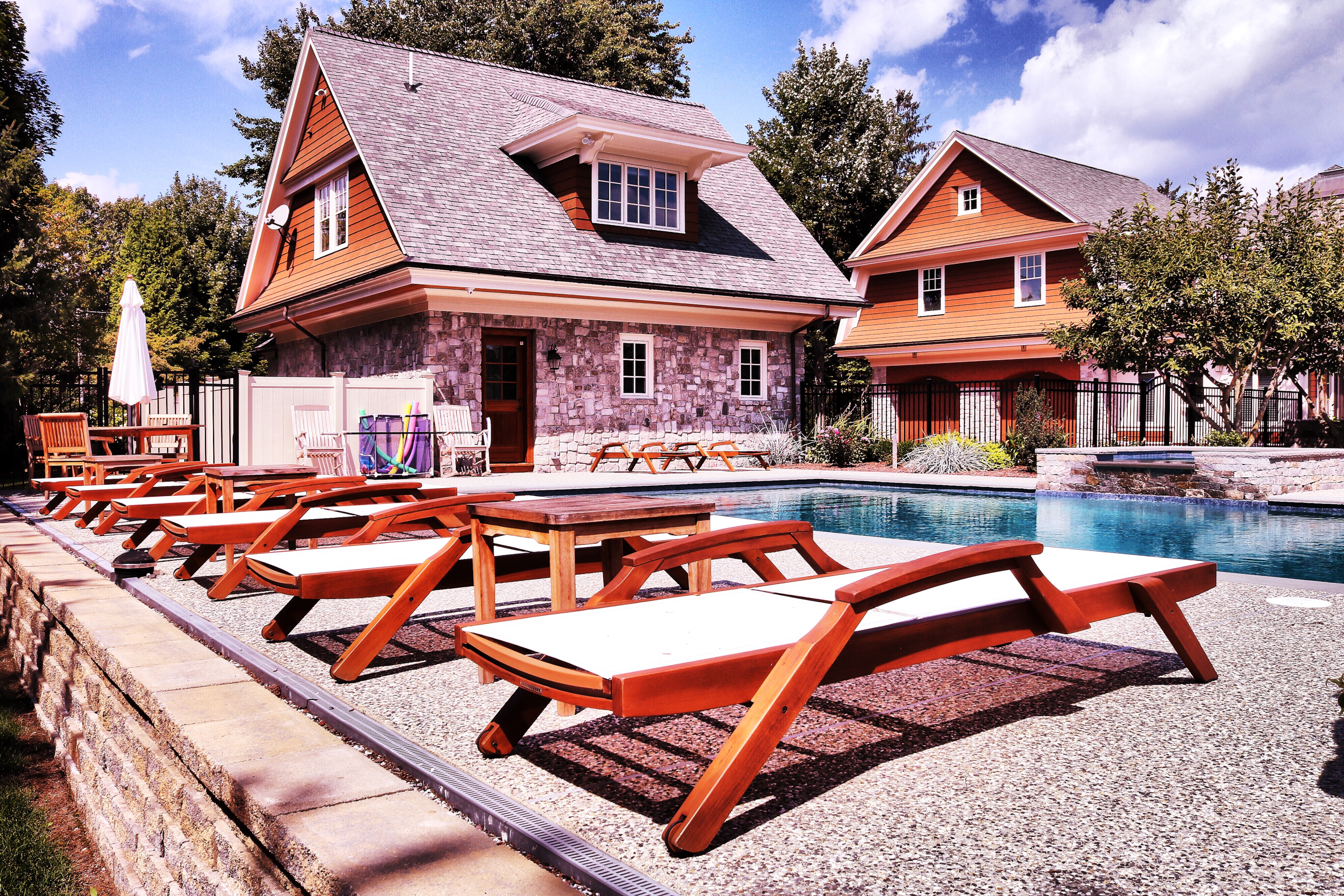 Outdoor swimming pool area with wooden lounge chairs and surrounding trees, next to a stone house and adjacent building under a partly cloudy sky.