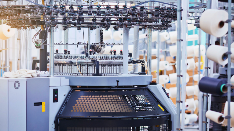 A close-up of a large industrial knitting machine with many spools of white, black, and polyester thread in the background.