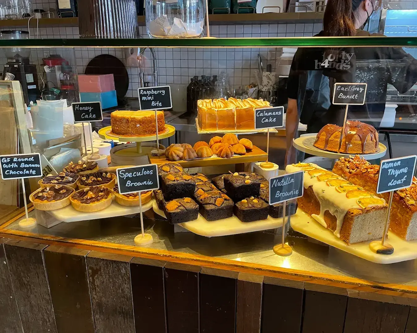 A bakery display with various cakes and brownies labeled, including Honey Cake, Earl Grey Bundt, and Nutella Brownie. A cozy, inviting atmosphere.