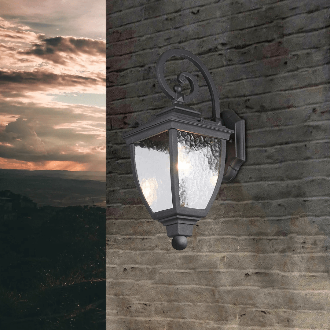 Black outdoor wall lantern with frosted glass panels and decorative scroll design, mounted on a brick wall at sunset.