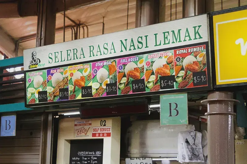 An illuminated menu signboard for the Selera Rasa Nasi Lemak food stall, displaying colorful photos and prices of various traditional meal combinations.