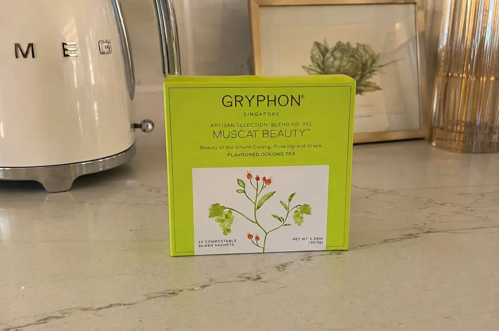 A square, bright lime-green box of Gryphon Singapore's Artisan Selection Blend No. 761, "Muscat Beauty," rests centrally on a modern, speckled white quartz kitchen countertop. The front of the box displays a white rectangular block featuring a detailed botanical illustration of a green grapevine branch with small red berries, identifying the product as a flavoured oolong tea with 10 compostable silken sachets. To the left, a portion of a sleek, cream-colored retro Smeg electric kettle is visible, while a framed print of an artichoke and a fluted amber glass vase sit softly out of focus in the background.