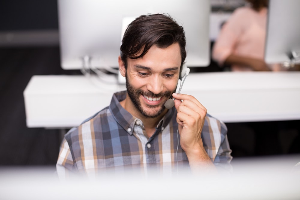  Smiling male customer service agent talking on headset at desk on a business phone.