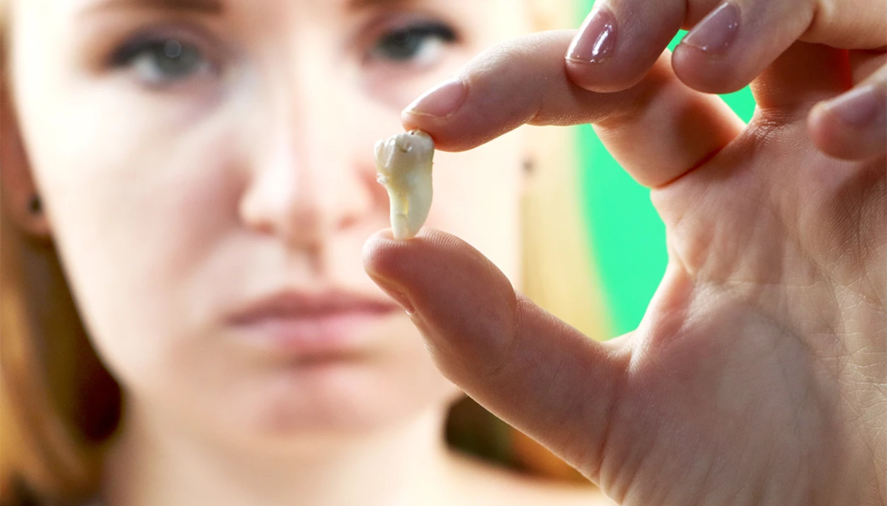 Kvinne holder en trukket tann mellom fingrene og ser alvorlig på den med uskarpt ansikt i bakgrunnen.