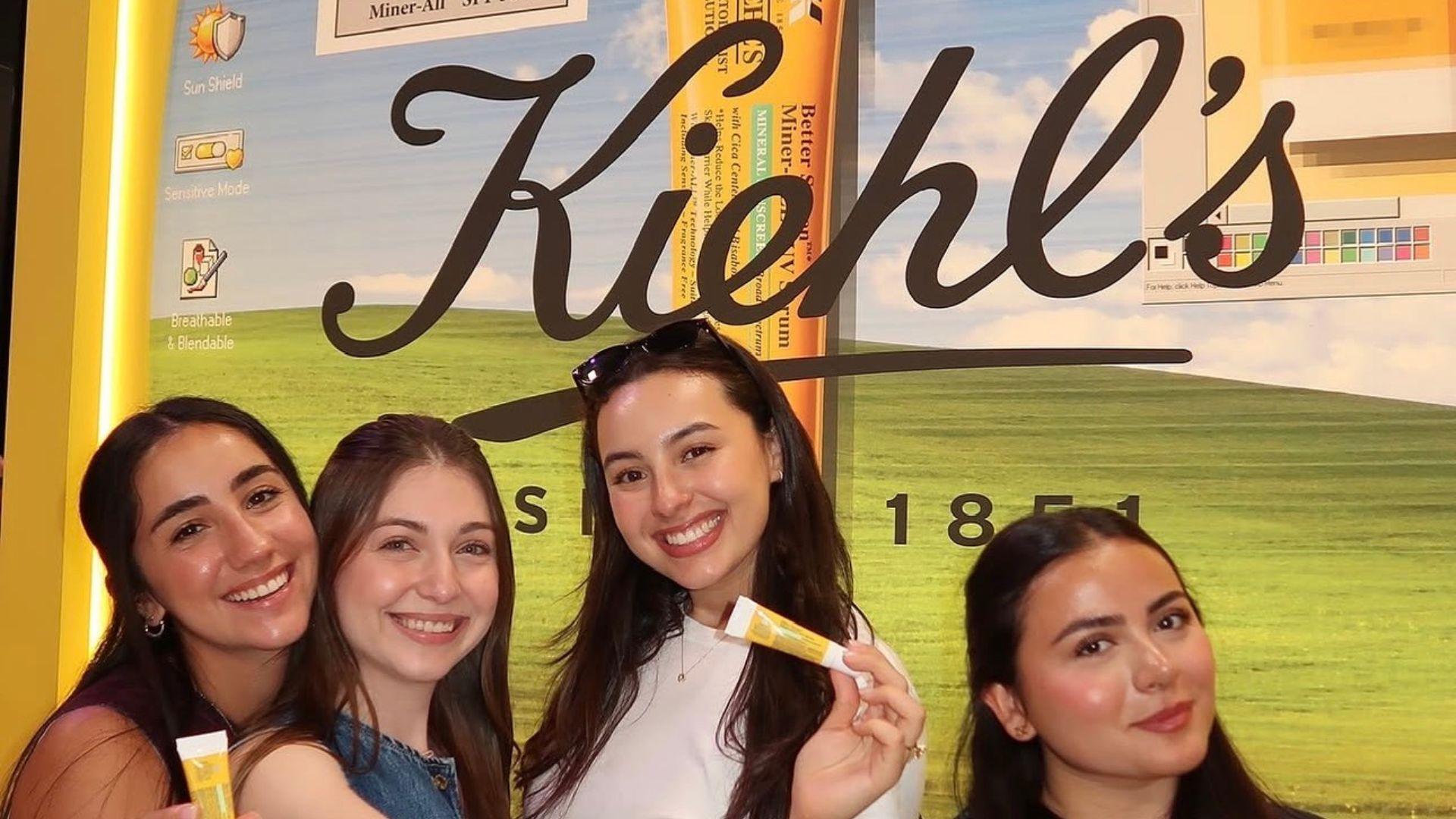 Four girls are smiling in front of a sign that says Kiehl's holding a lip product.