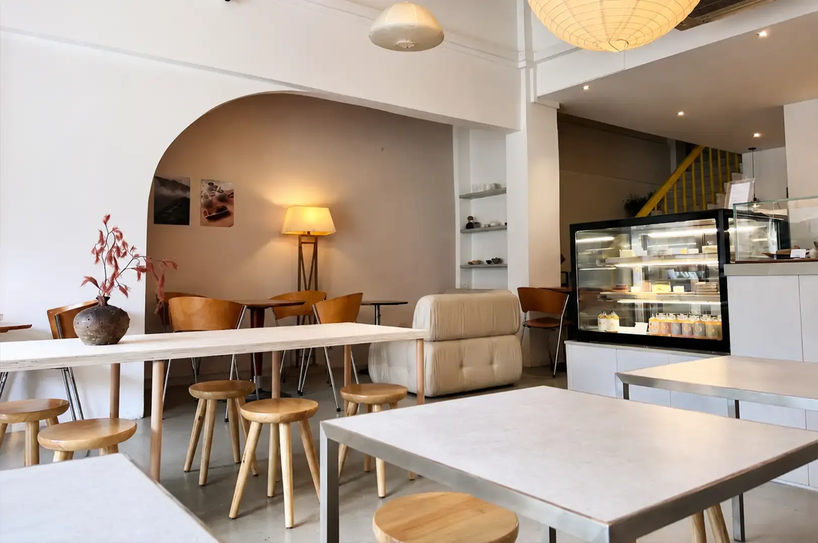 An eye-level view shows the airy interior of the teahouse, characterized by white walls, polished concrete floors, and light wooden furniture. Light-colored wooden tables paired with round three-legged stools and bentwood chairs fill the space, while an oversized paper lantern hangs from the high ceiling. In the background, a warm floor lamp illuminates an arched seating nook, adjacent to a glass display case stocked with cakes and chilled bottled drinks.
