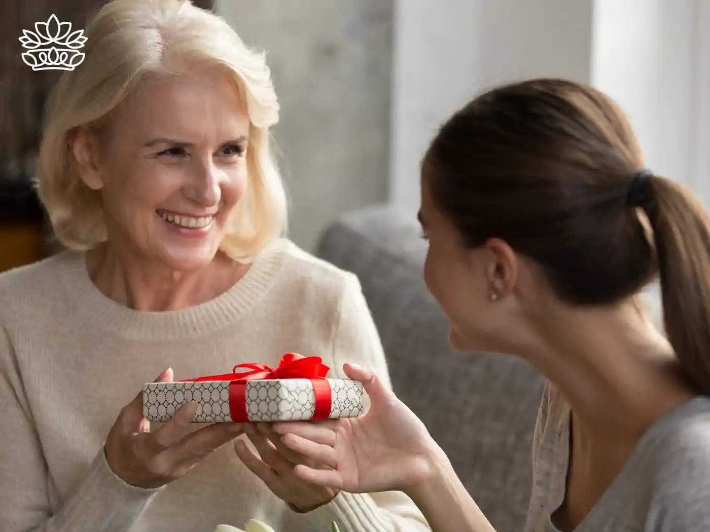 Young woman giving her elderly mother a beautifully wrapped gift box, both smiling warmly in a cozy indoor setting. 