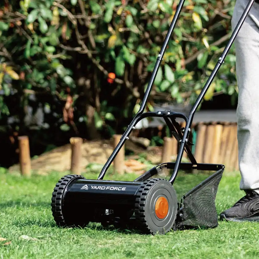 A person is pushing a Yard Force manual lawn mower on a green lawn, with a lush garden and wooden border in the background. The mower's design is sleek and black, featuring a mesh grass catcher at the back.